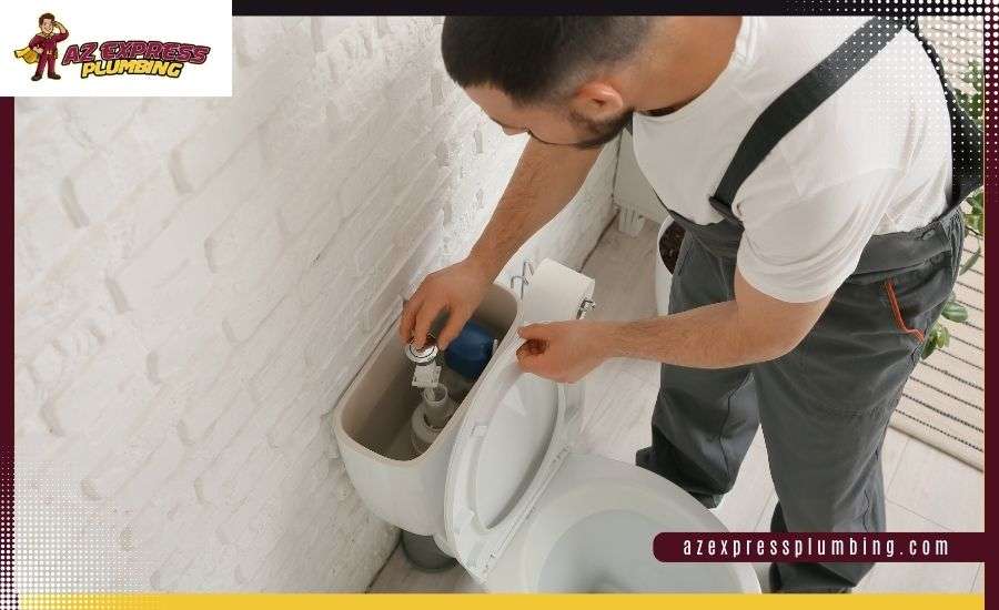 A plumber in grey overalls inspects the internal flushing mechanism of an open white toilet tank against a white brick wall.